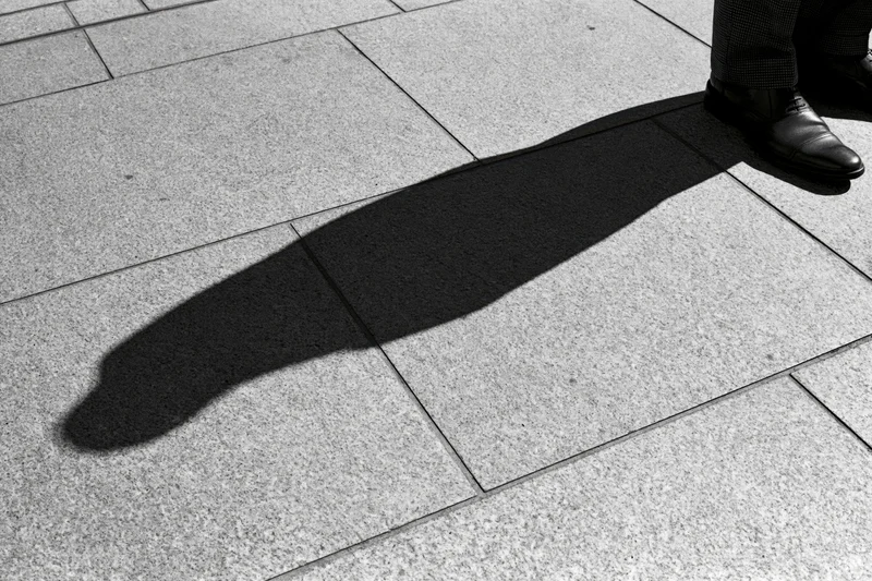 Long shadow of a standing person cast across grey pavement in urban setting
