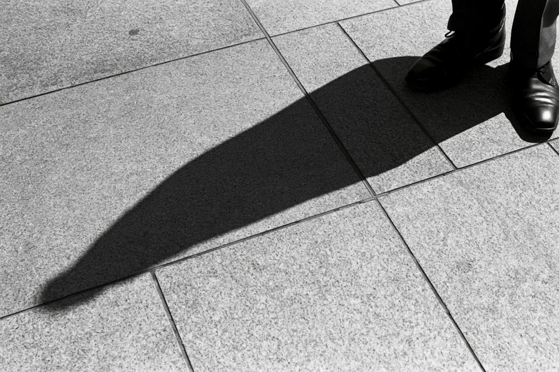 Long shadow of a standing person cast across grey pavement in urban setting