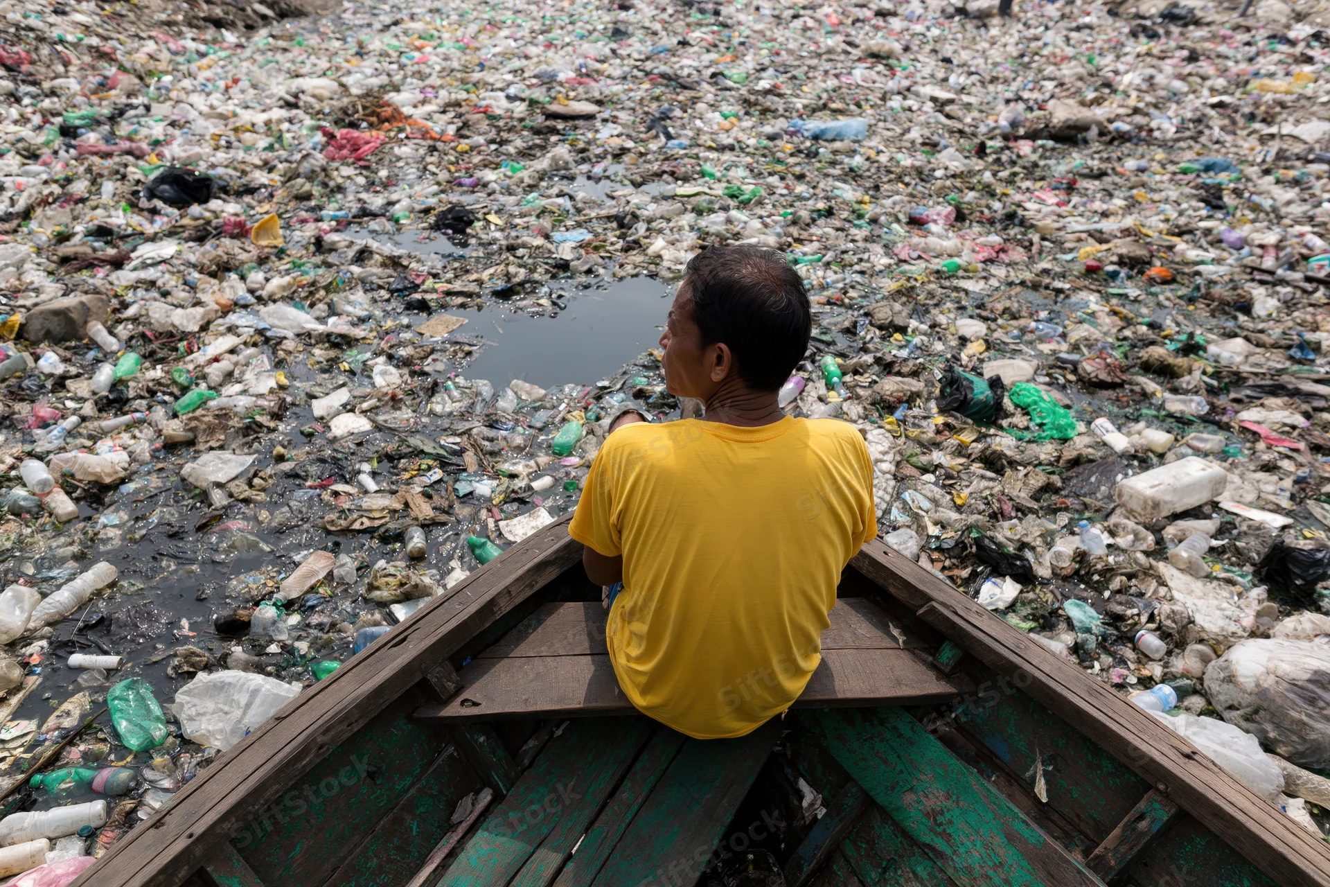 Man sitting at bow of wooden boat looking at plastic waste covering water surface