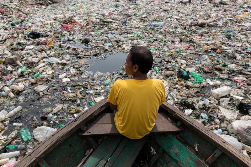 Man sitting at bow of wooden boat looking at plastic waste covering water surface