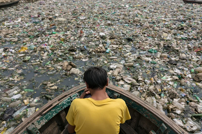 Man in wooden boat facing river choked with plastic waste