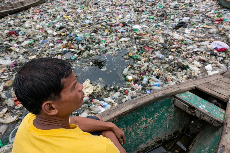 Man sitting at bow of weathered wooden boat looking at plastic waste floating in polluted river