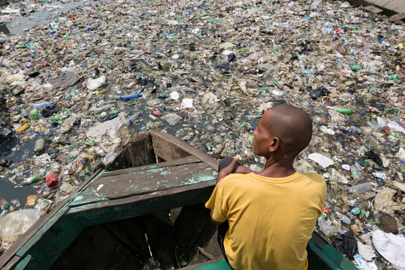 Man sitting at bow of weathered wooden boat looking at river filled with plastic waste