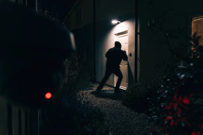 Masked figure in black breaking into a house at night, seen from behind a surveillance camera with red light