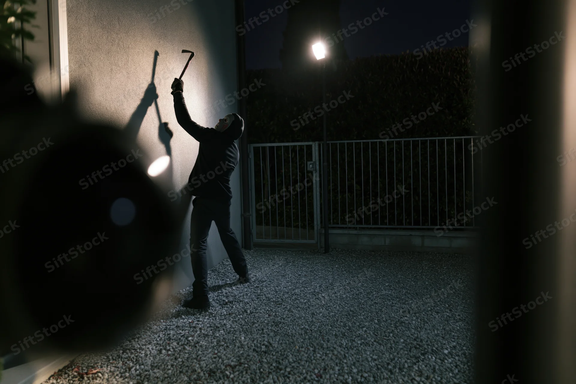 Hooded burglar raising crowbar against wall at night near gate