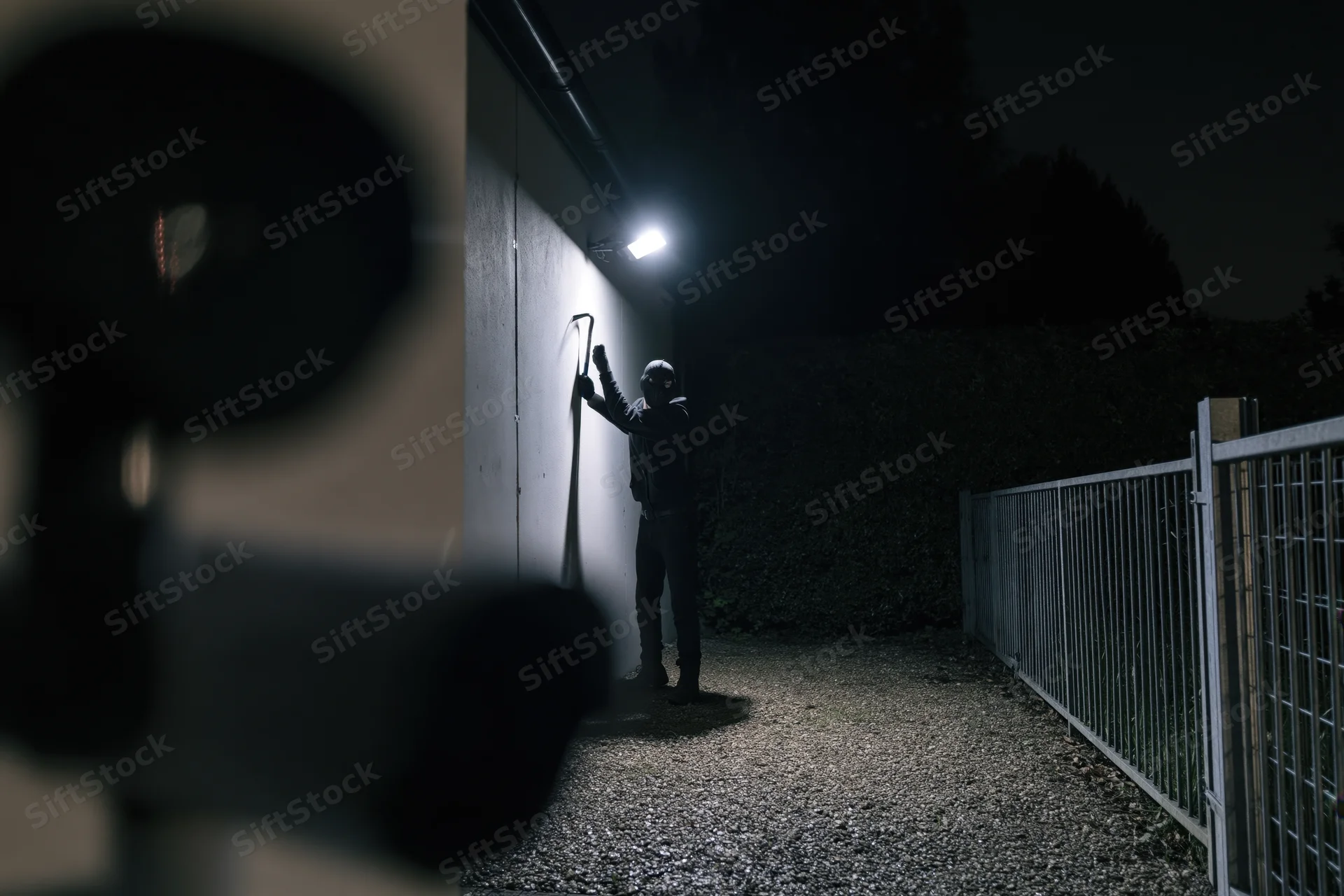 Masked burglar raising crowbar against wall in dark alley at night, viewed through blurred camera lens