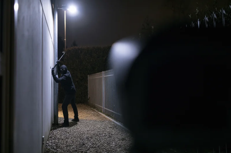 Masked burglar using crowbar to pry open a wall at night in a residential alley