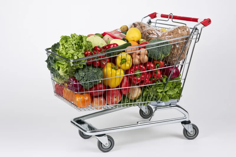 Metal shopping cart filled with fresh vegetables and fruits on white background