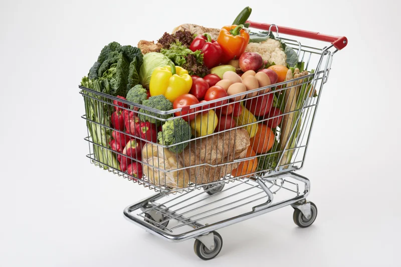 Metal shopping cart filled with fresh vegetables, fruits, eggs and bread on white background