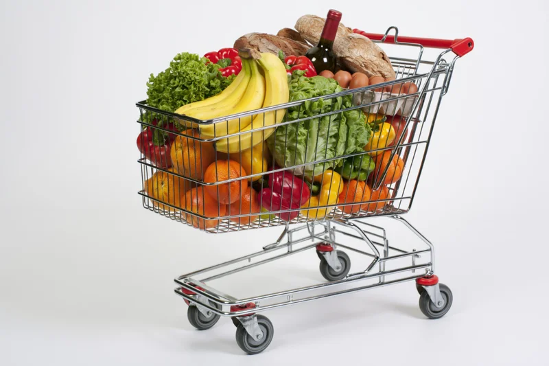 Metal wire shopping cart filled with fresh fruits vegetables bread eggs and wine on white background