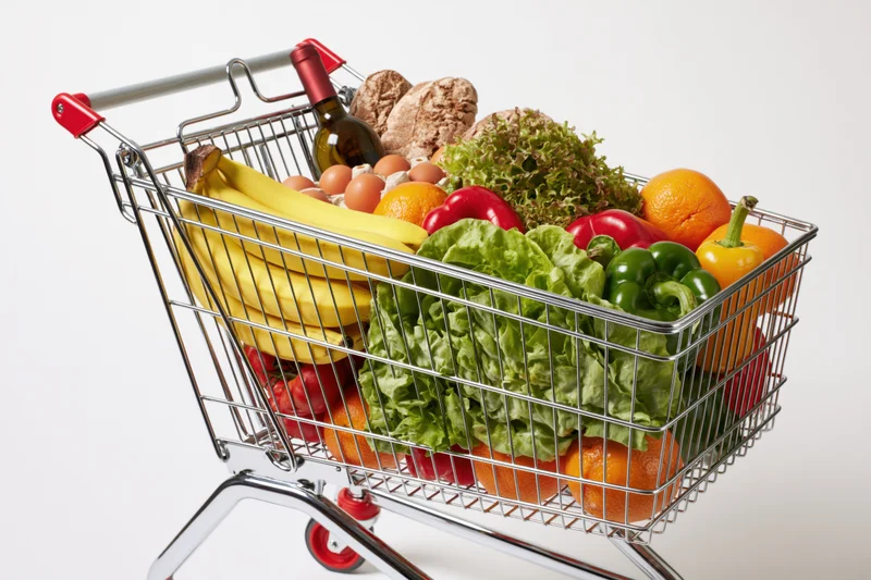 Metal wire shopping cart filled with fresh fruits vegetables bread eggs and wine on white background