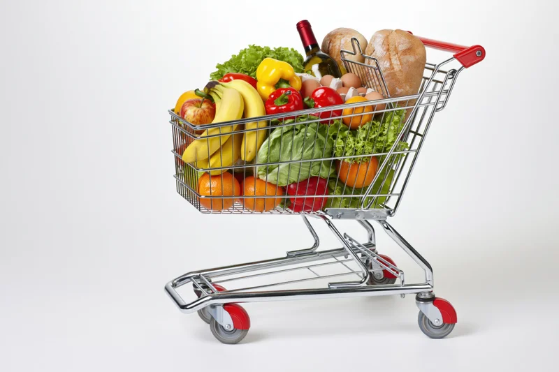 Metal wire shopping cart filled with fresh fruits vegetables bread eggs and wine on white background