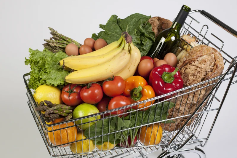 Metal wire shopping cart overflowing with fresh produce and groceries on white background