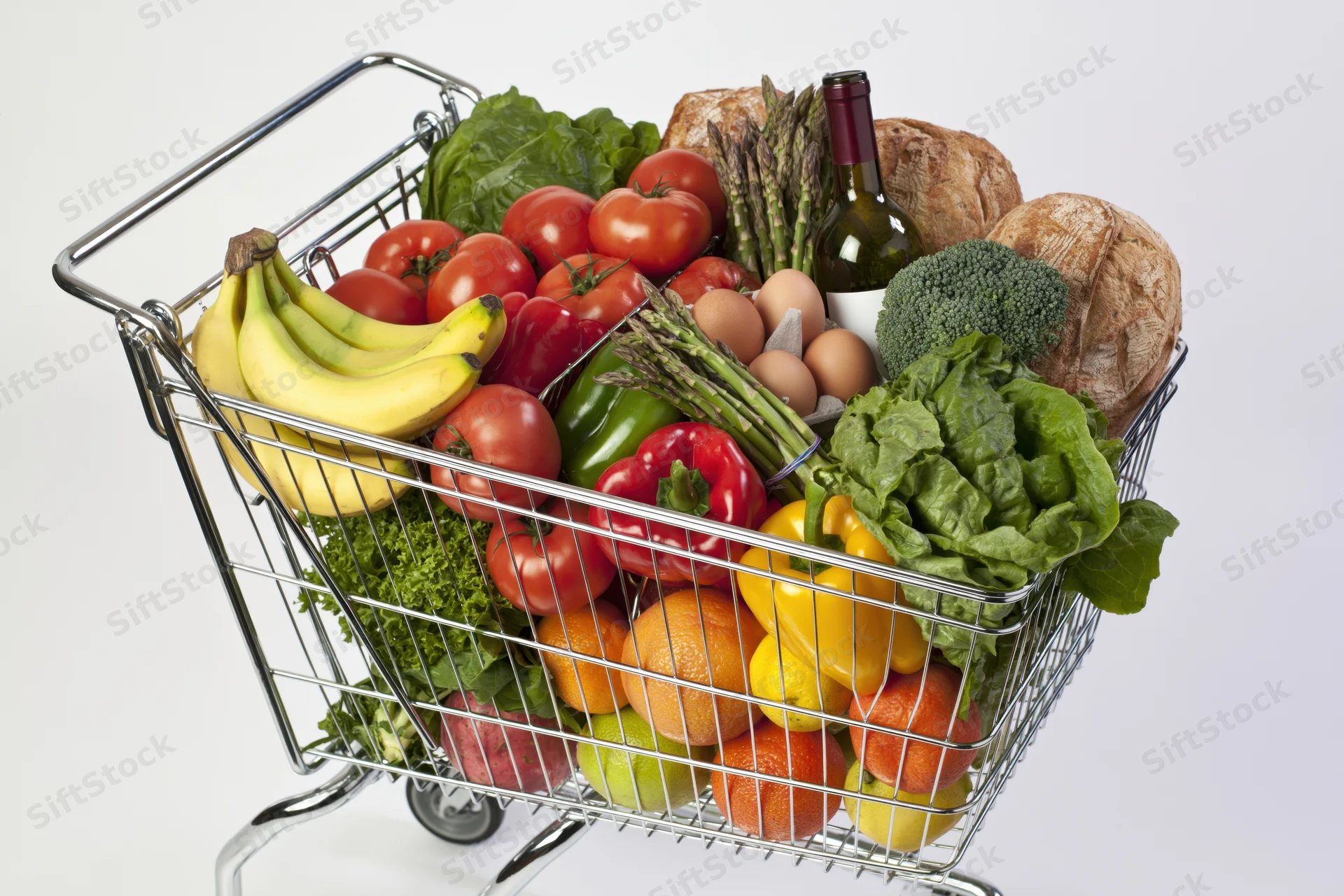 Metal wire shopping cart overflowing with fresh fruits vegetables bread and wine against white background