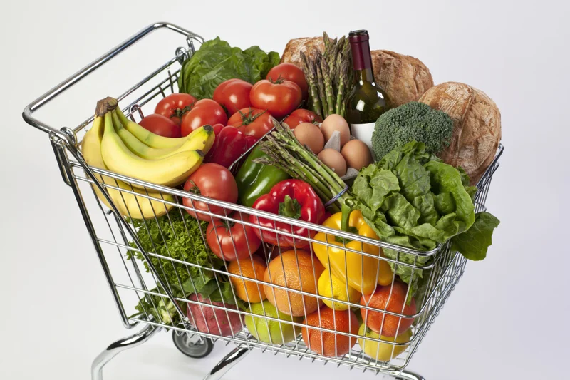 Metal wire shopping cart overflowing with fresh fruits vegetables bread and wine against white background