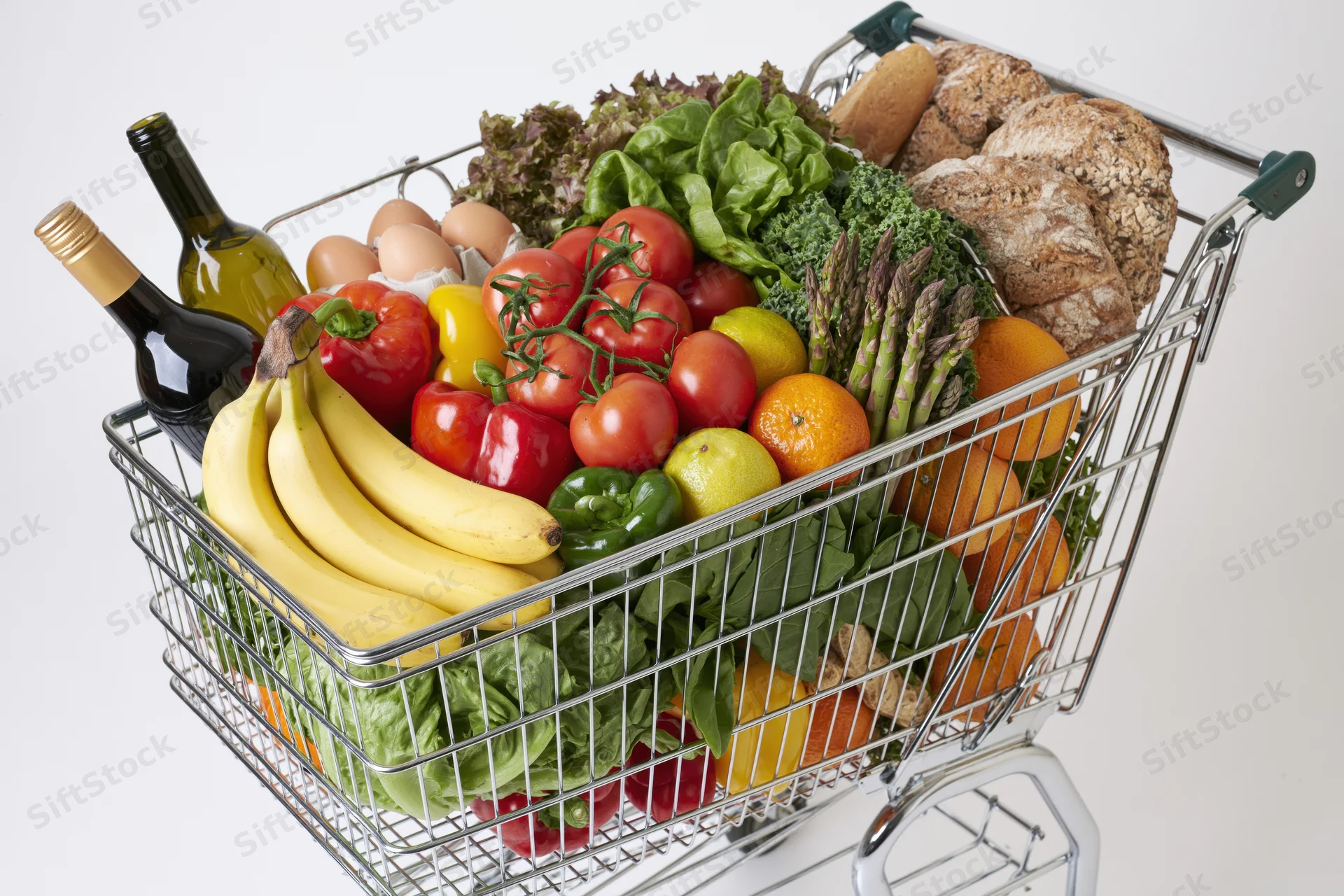 Metal wire shopping cart overflowing with fresh produce and groceries against white background