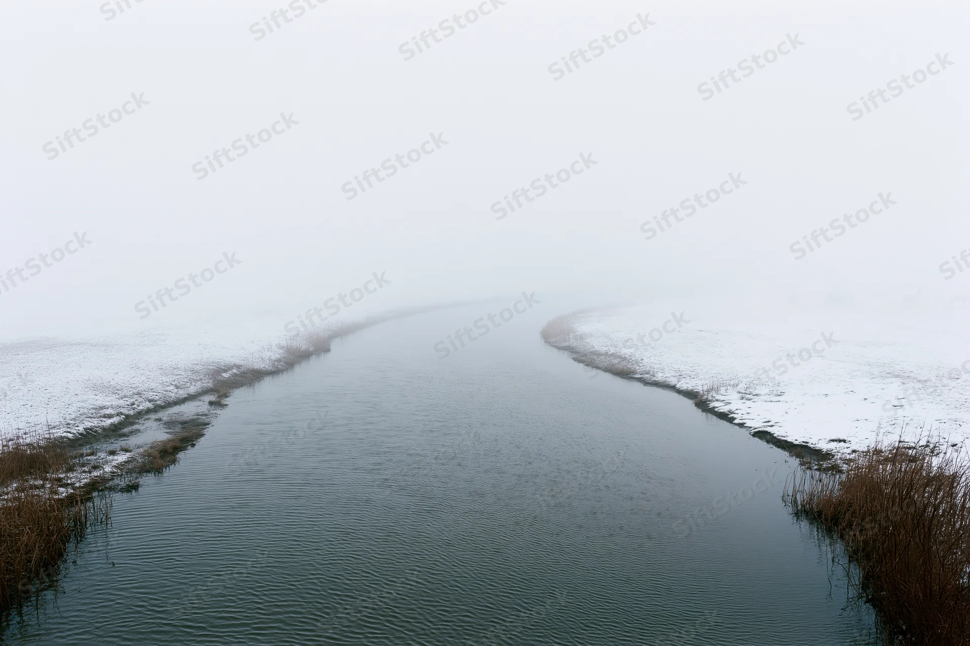 Narrow winding river flowing through snow-covered banks into foggy distance