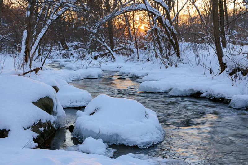 Partially frozen river flowing through snow-covered forest at sunset