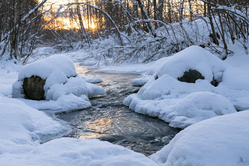 A partially frozen river flows between snow-covered rocks and trees at sunset