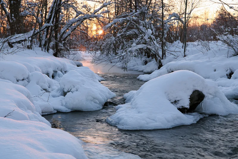 A partially frozen river flows between snow-covered banks at sunset in a forested winter landscape