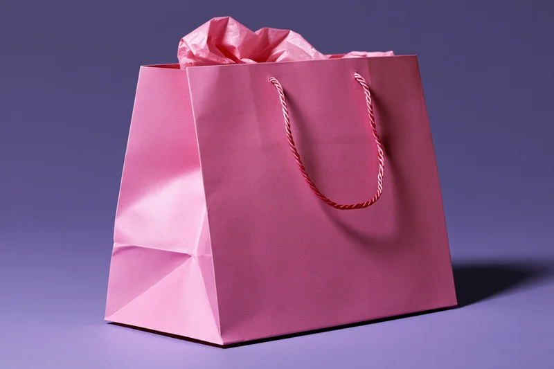 Pink paper shopping bag with crumpled tissue paper inside, tilting on purple surface