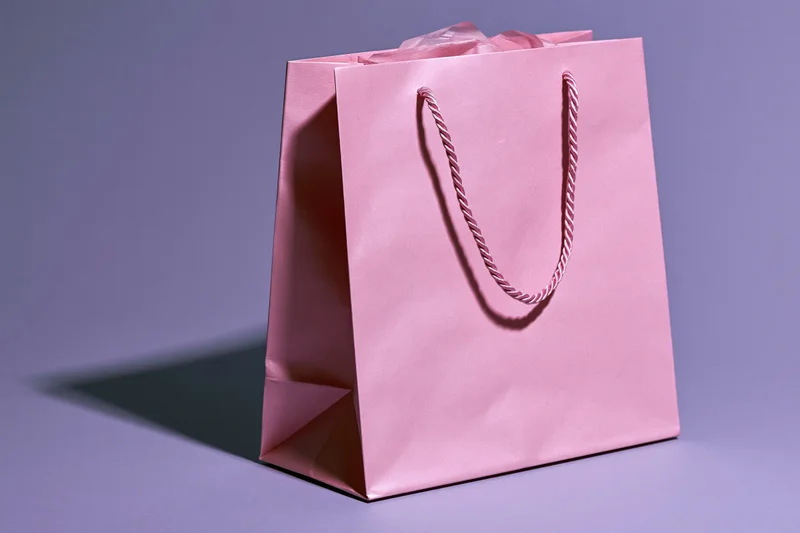 Pink paper shopping bag standing at an angle on a purple surface with twisted cord handles