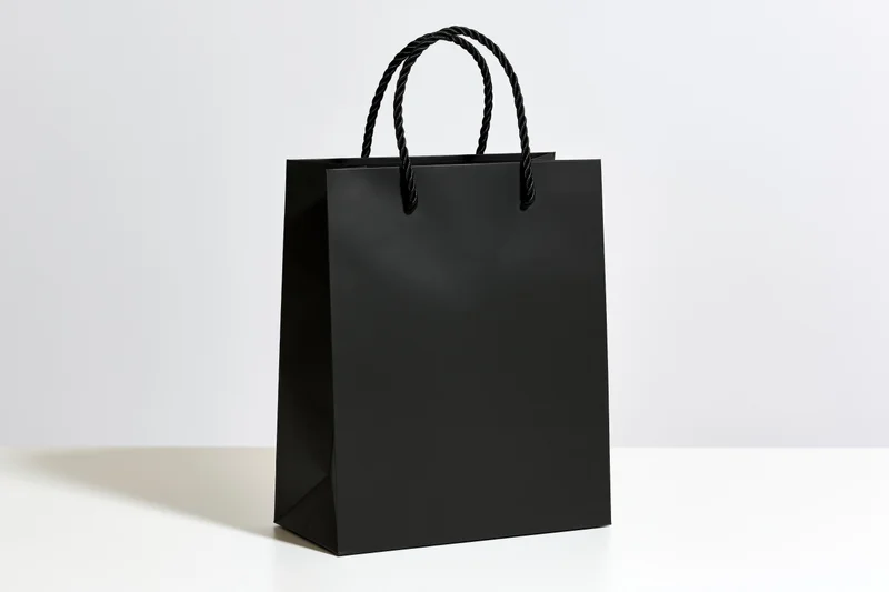 Plain matte black paper shopping bag with twisted rope handles on white surface