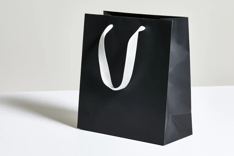 Matte black paper shopping bag with white ribbon handles on white surface