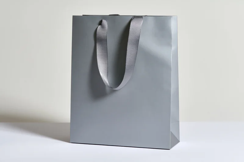 Matte grey paper shopping bag with woven ribbon handles standing on white surface