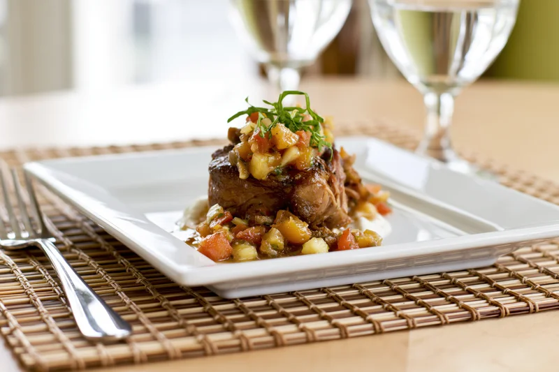 Braised meat dish with vegetable sauce on a white square plate set on a bamboo placemat with fork and wine glasses nearby