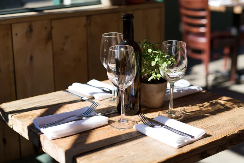 Wooden restaurant table set for two with empty wine glasses and cutlery under sunlight