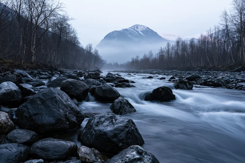 River flowing over boulders in a misty mountain valley surrounded by bare trees and snow-capped peaks