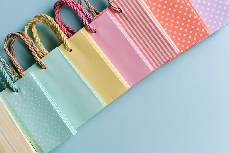 Row of colorful paper shopping bags with rope handles arranged diagonally on light blue surface