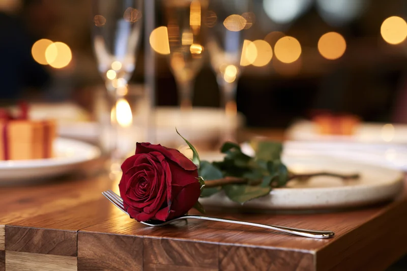 Deep red rose resting on silver fork on wooden table at romantic dinner setting