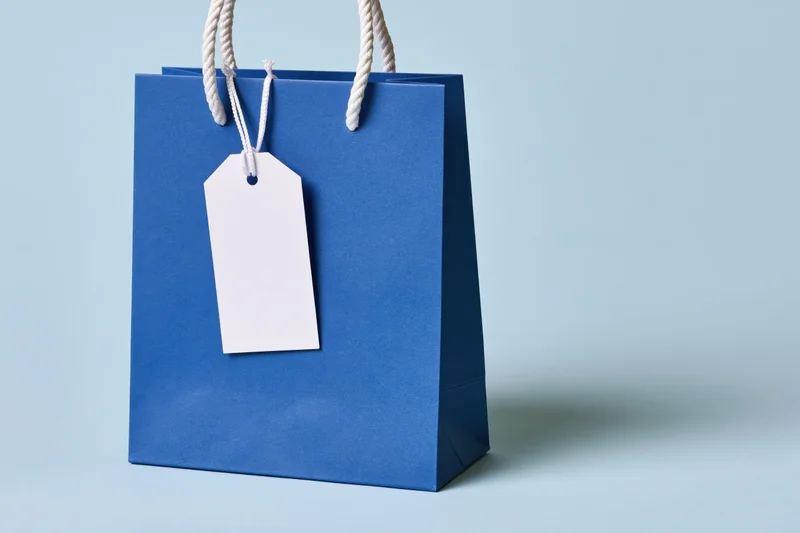 Cobalt blue paper shopping bag with white rope handles and blank tag on light background