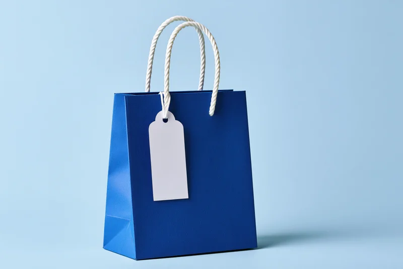 Cobalt blue paper shopping bag with white rope handle and blank tag on light blue background