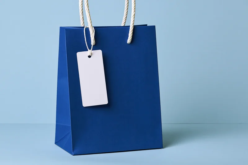 Cobalt blue paper shopping bag with white rope handle and blank tag on light blue surface
