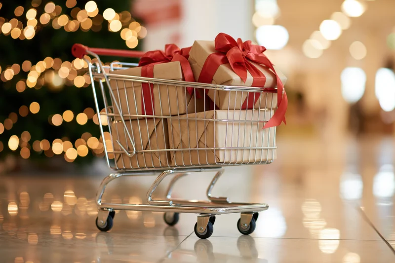 Small metal shopping cart filled with wrapped gifts in a mall with blurred holiday lights in the background