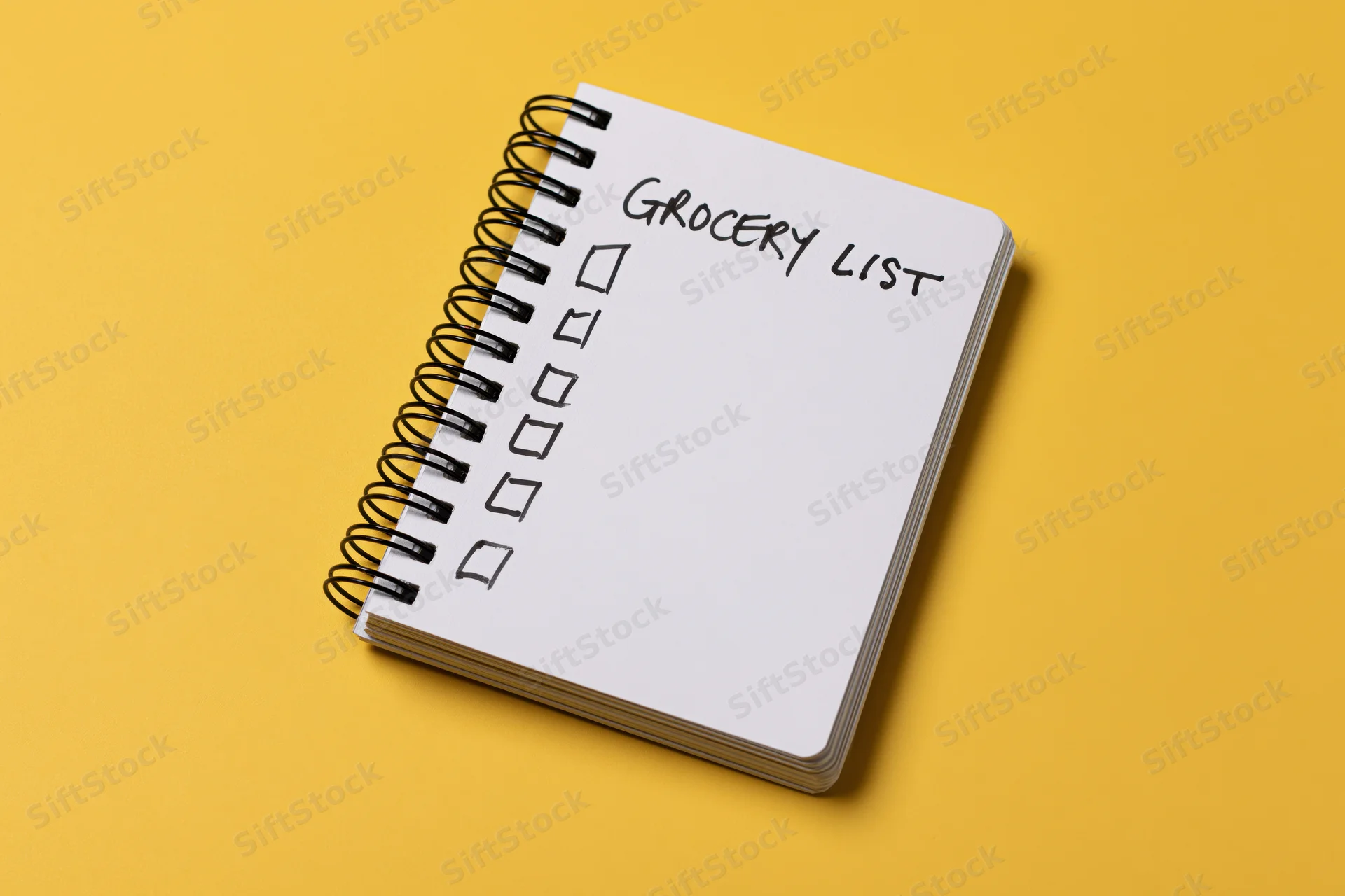 Spiral-bound notepad with handwritten grocery list and checkboxes on yellow background