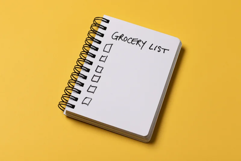 Spiral-bound notepad with handwritten grocery list and checkboxes on yellow background
