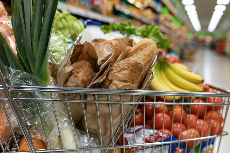 Shopping cart filled with baguettes, tomatoes, bananas, green onions and milk in a supermarket aisle