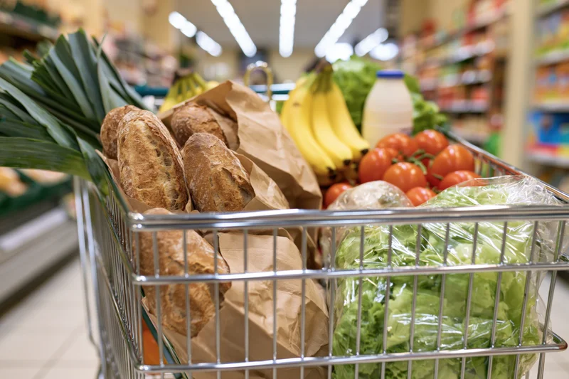 Shopping cart filled with baguettes, bananas, tomatoes, lettuce and milk in a supermarket aisle
