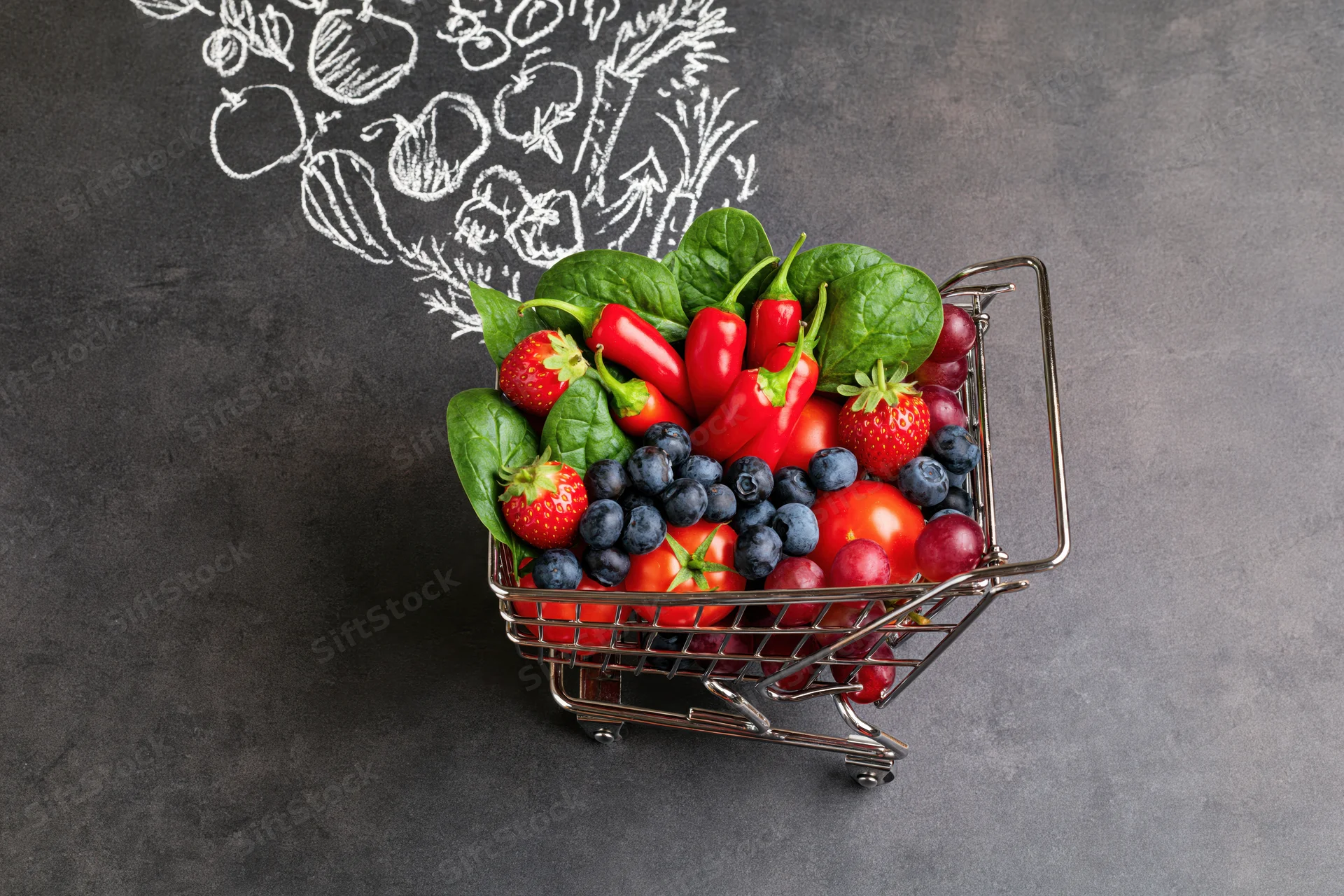 Tiny metal shopping cart filled with fresh fruits and vegetables on dark surface with chalk vegetable sketch behind