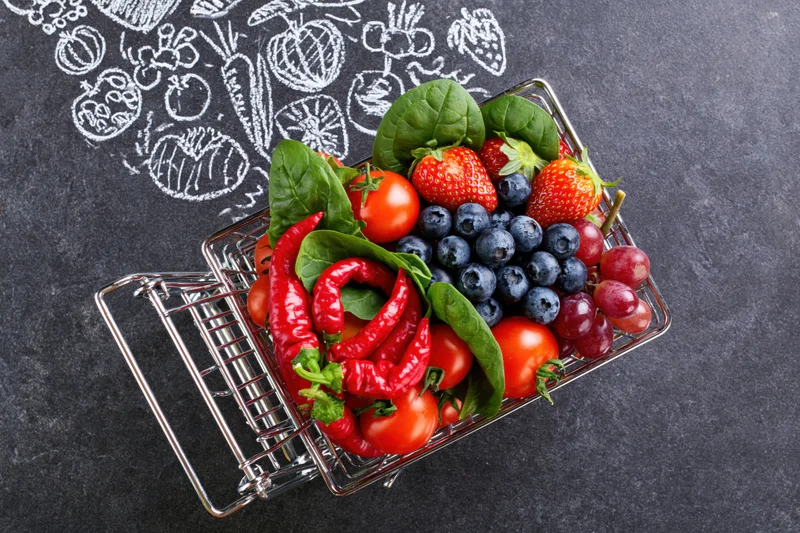 Tiny metal shopping cart filled with fresh fruits and vegetables on dark surface with chalk vegetable drawings