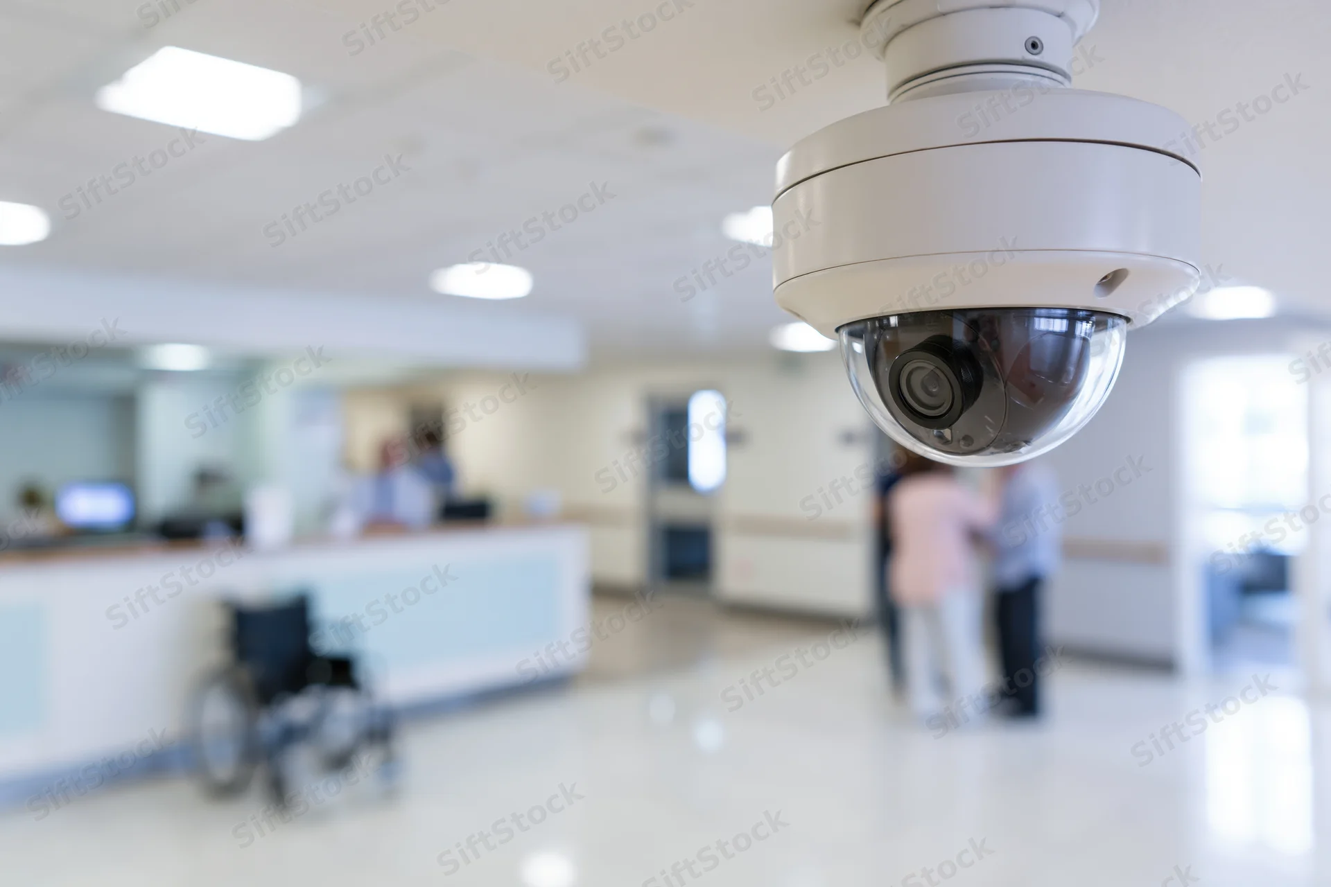 White dome security camera mounted on ceiling in hospital lobby with reception desk and wheelchair in background