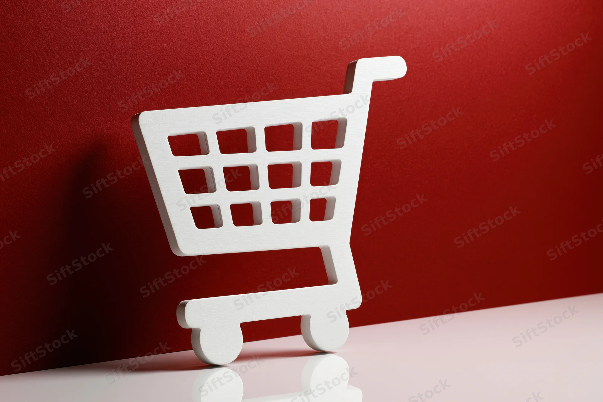 White shopping cart icon prop standing upright on a red background with reflection