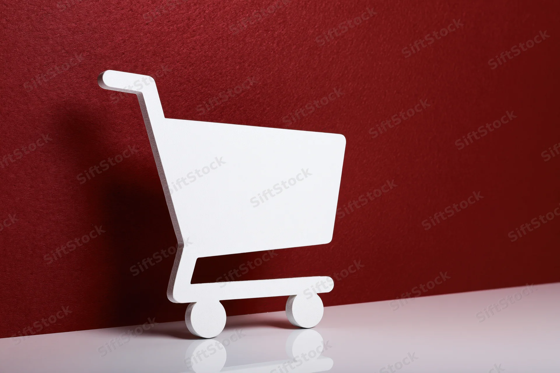 White flat shopping cart icon prop standing upright on a red textured wall with reflective white surface