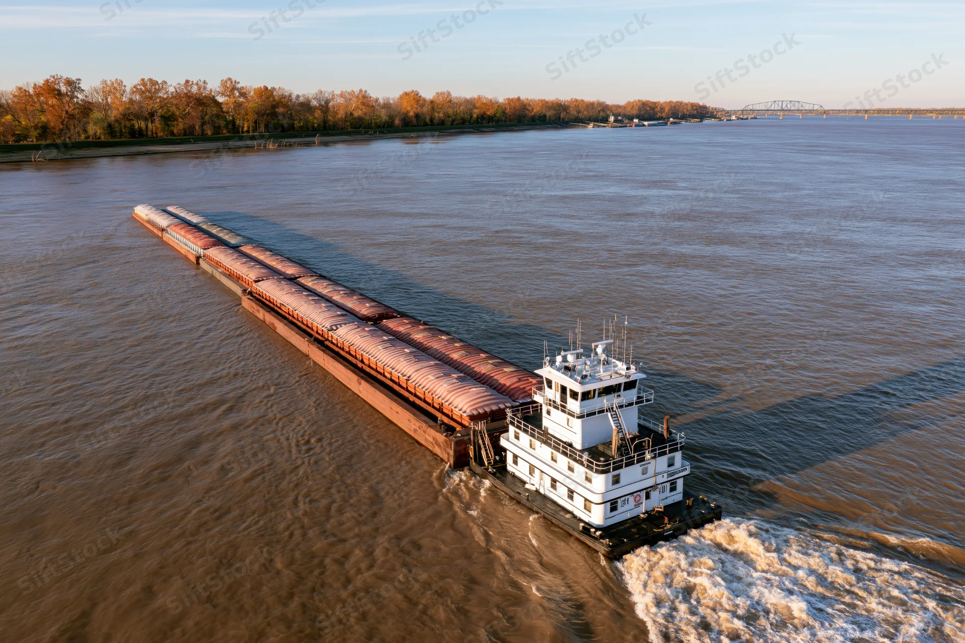 White towboat pushing long string of covered cargo barges on wide river at golden hour