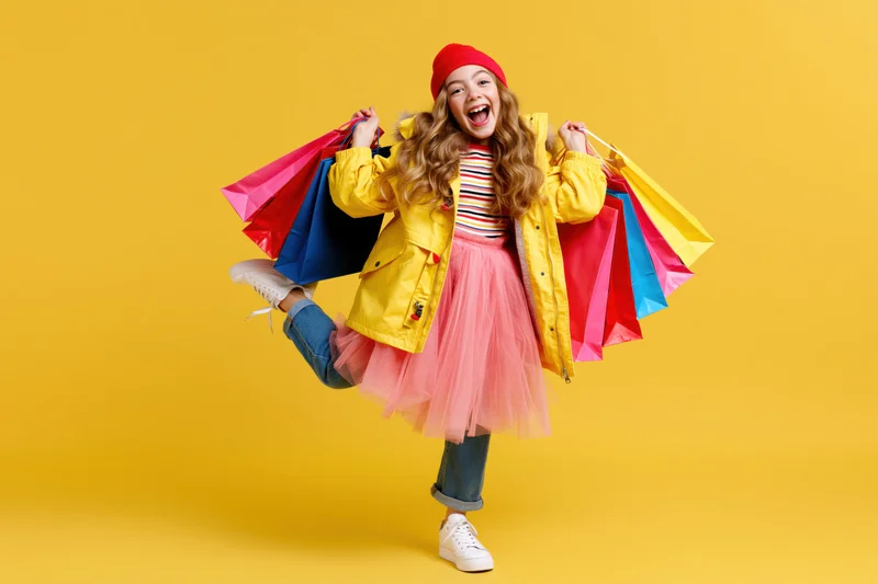 Young girl laughing and kicking one leg while holding colorful shopping bags against yellow background