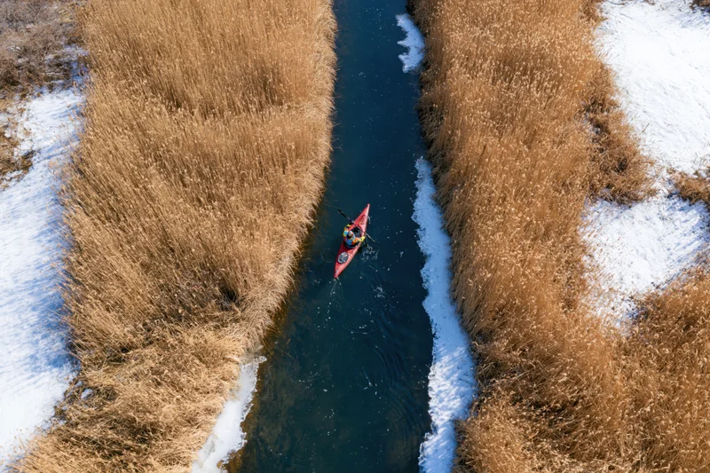 Aerial view of a man kayaking in a narrow river surrounded by snow and tall reeds